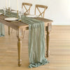 A rustic table set featuring a green cotton gauze table runner on a wooden table with glassware and plates.