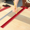 A hand is using a yellow pencil to mark a precision red aluminum alloy T-ruler on a wooden surface for woodworking.