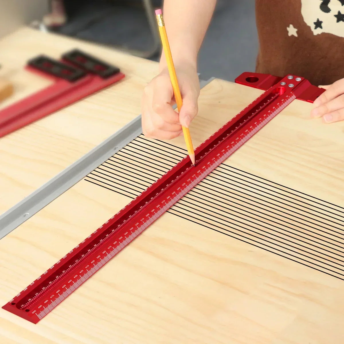 A hand is using a yellow pencil to mark a precision red aluminum alloy T-ruler on a wooden surface for woodworking.