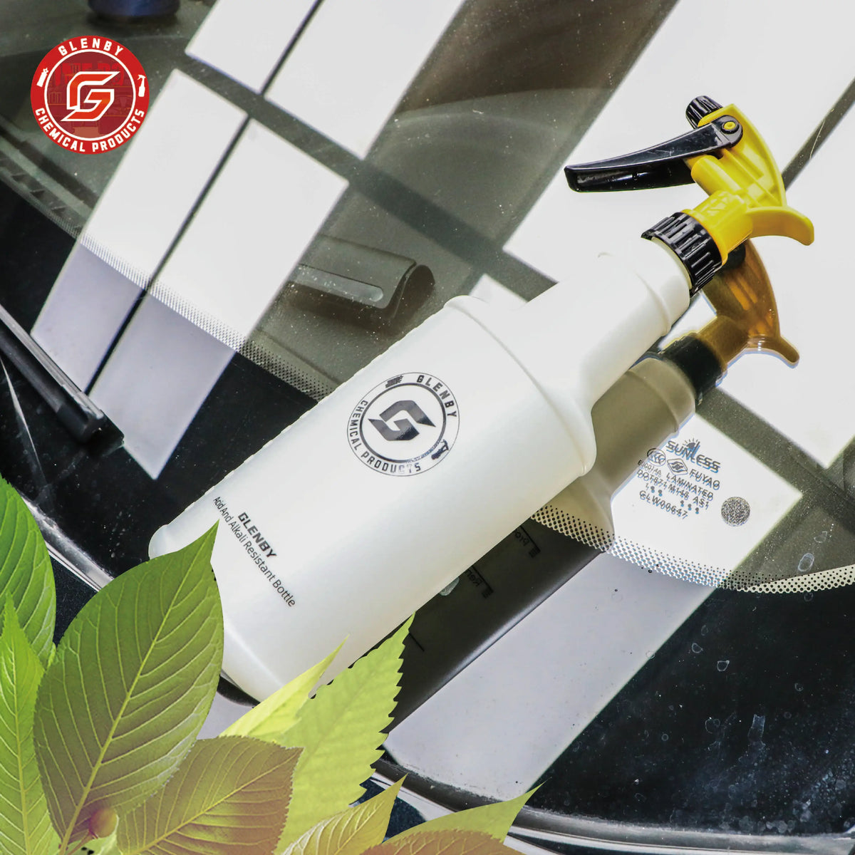 A white plastic spray bottle with a yellow nozzle is shown on a glass surface next to green leaves.