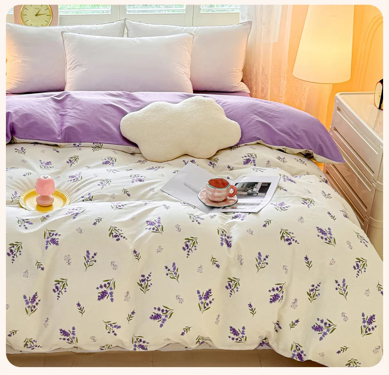 Lavendelblüchse-Kopftepp, weiße Akzente und eine cremefarbene Wolkenkissen auf einem Bett.