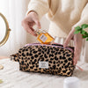 A hand is pouring oil from a glass bottle into a leopard print toiletry bag on a white surface.