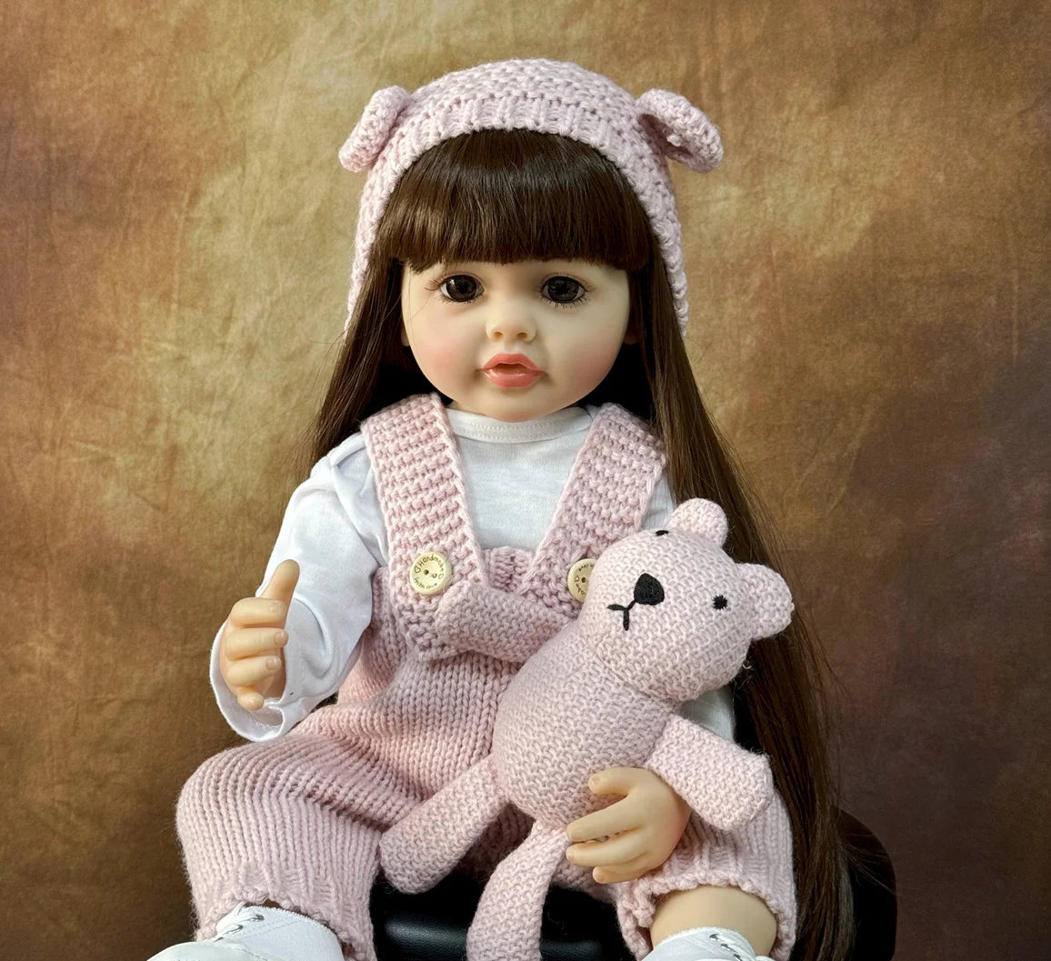 A lifelike silicone baby doll wearing a pink knitted hat and overalls, holding a small pink stuffed animal.