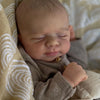 A newborn baby doll with closed eyes resting on a soft, light-colored fabric background.