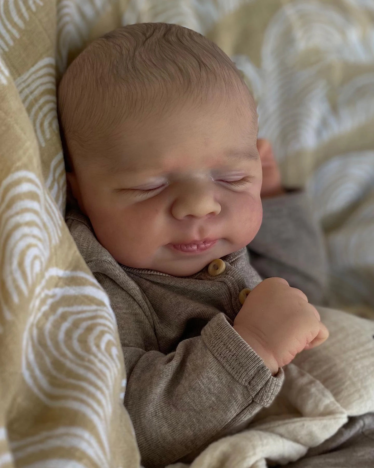 A newborn baby doll with closed eyes resting on a soft, light-colored fabric background.