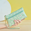 A hand holds a light green daisy print PU leather wristlet wallet with a zipper closure against a yellow background.