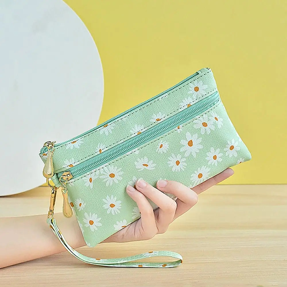 A hand holds a light green daisy print PU leather wristlet wallet with a zipper closure against a yellow background.