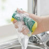 A hand is holding a green and yellow double-sided cleaning sponge under running water in a kitchen sink.