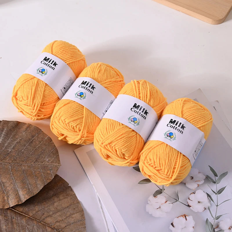Several balls of yellow Milk Cotton yarn are displayed on a white surface next to a dried leaf and small white flowers.