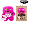 A bright pink Fuggler Ugly Monster plush toy with large teeth and a white bib, packaged in a cardboard box.
