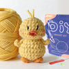 A yellow crocheted bird toy next to a purple bag of crochet kit supplies and a needle.