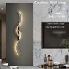 A modern minimalist LED wall lamp with a double S shape is mounted on a textured gray wall in a bright interior.