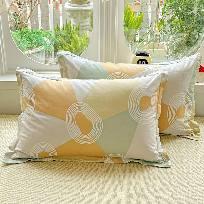 A set of light yellow, white, and sage green cotton pillow shams displayed in front of a window with greenery.