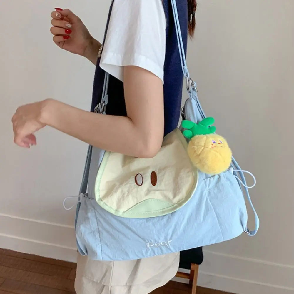 A person is holding a light blue nylon messenger bag with a yellow embroidered face and green handles.