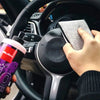 A hand is using a gray microfiber scrub sponge to clean the steering wheel of a car.