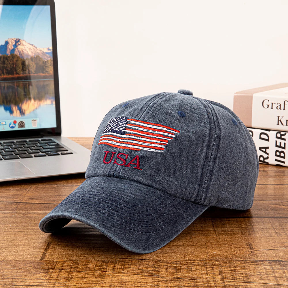 A dark gray baseball cap embroidered with the American flag design resting on a wooden surface next to a laptop.