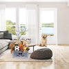 A gray felt pet toy storage basket with wooden handles sits on a rug in a bright room.