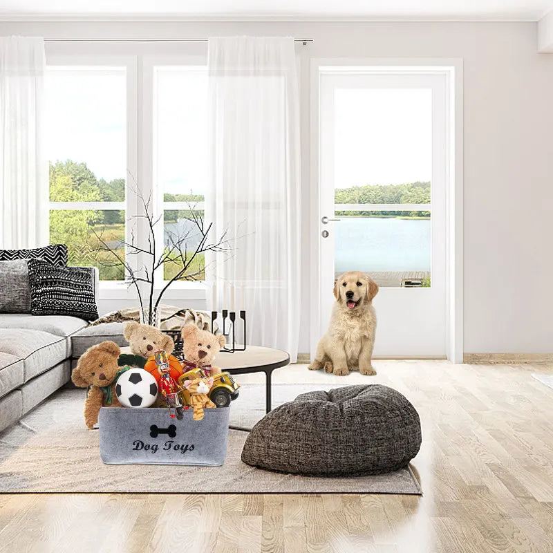 A gray felt pet toy storage basket with wooden handles sits on a rug in a bright room.