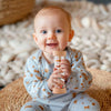 A smiling baby holding a wooden rain rattle toy in a light blue outfit, sitting on a woven mat.