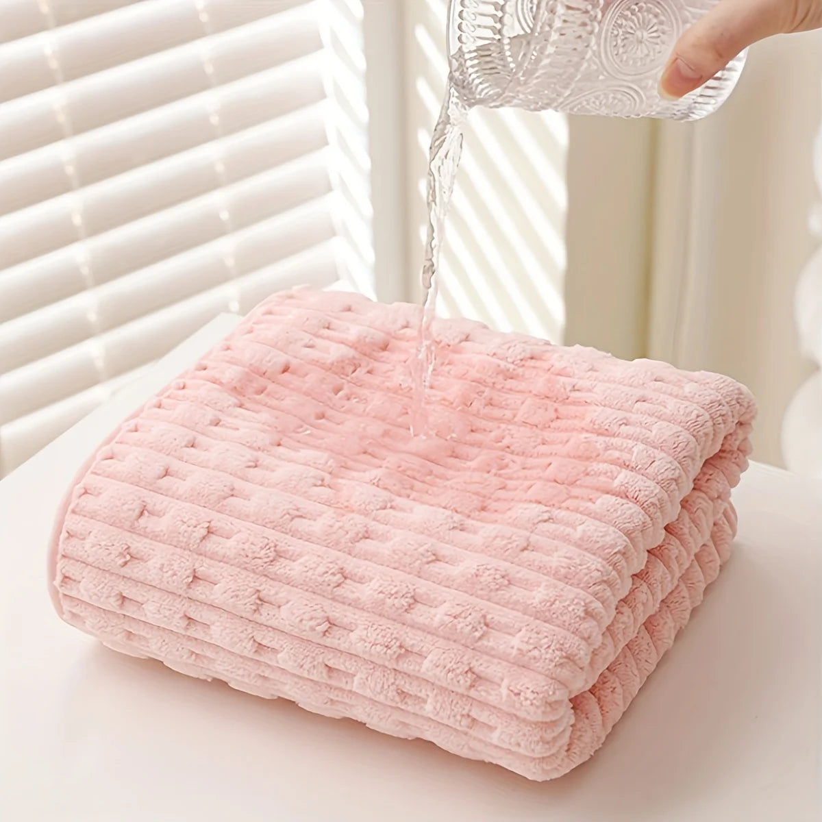 A pink coral velvet bath towel is being filled with water from a clear glass pitcher.