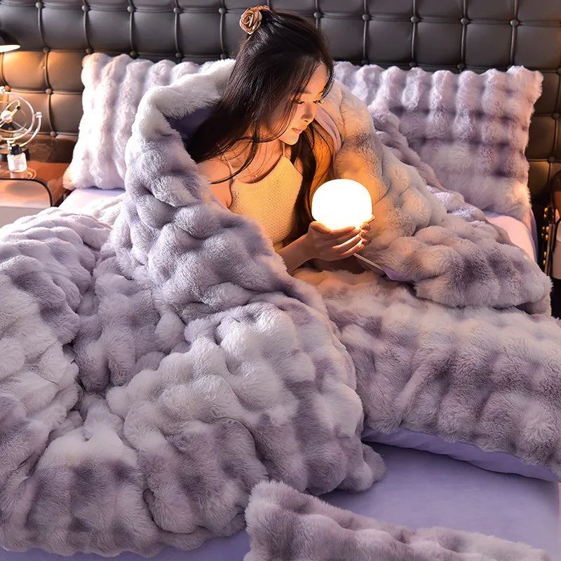 A woman is relaxing under a fluffy gray faux fur blanket on a bed.