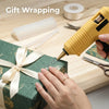 A hand uses a yellow hot glue gun to apply clear glue to a gift box for wrapping.