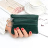 A hand holds a dark green compact PU leather two-zip coin purse next to a white mug and some papers.