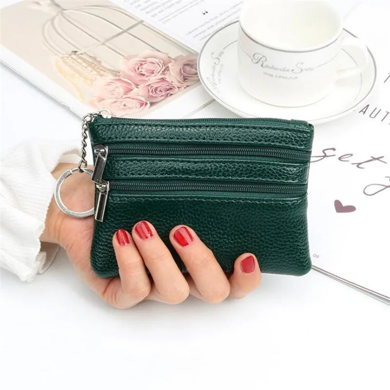 A hand holds a dark green compact PU leather two-zip coin purse next to a white mug and some papers.