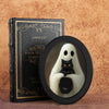 A black box with gold lettering sits next to an oval frame containing a white ghost figure and a black cat.