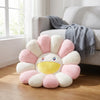 A cheerful sunflower plush cushion with a smiling face in pink, white, and yellow colors sits on a wooden floor.