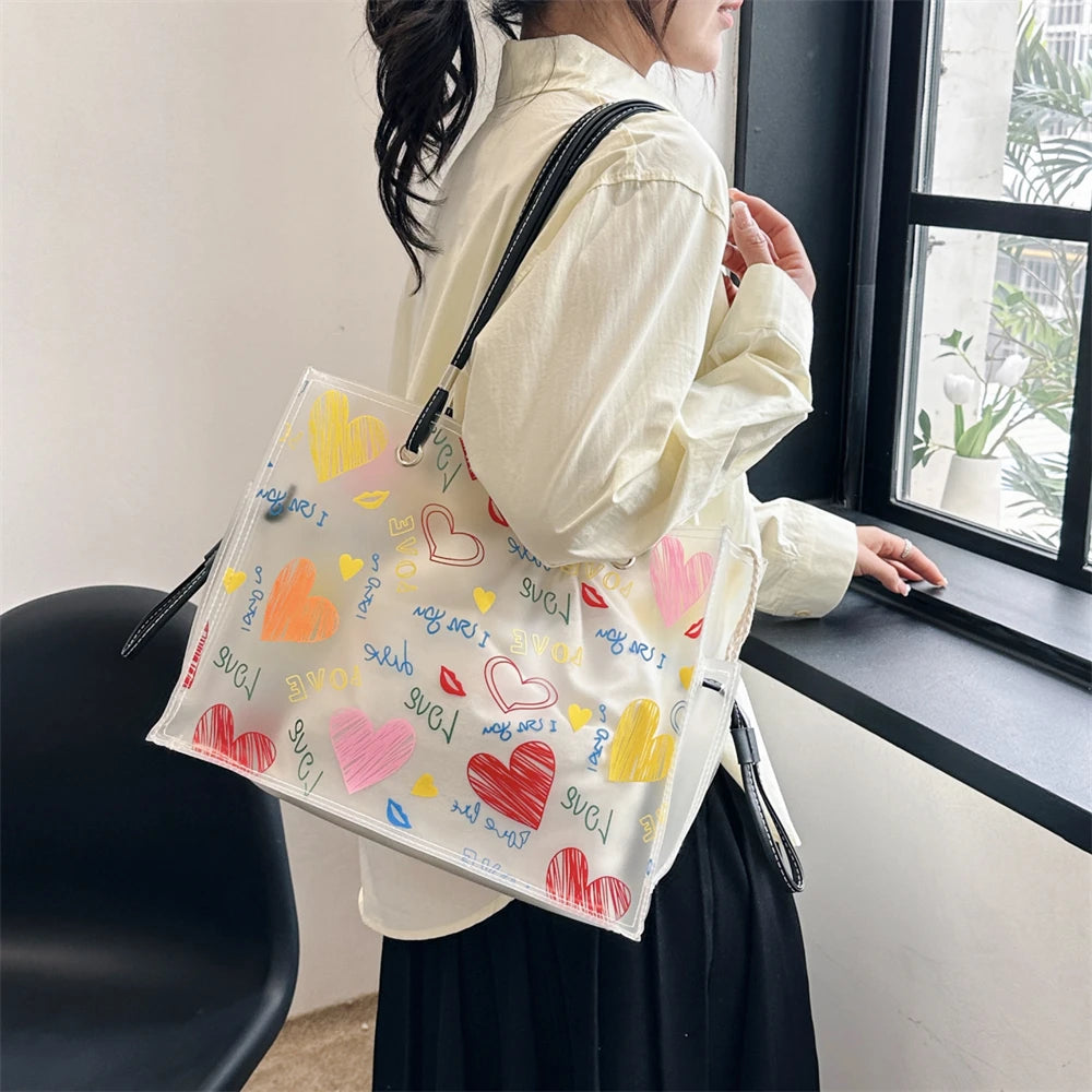 A woman holds a transparent tote bag with heart-shaped designs and an inner pouch next to a window.