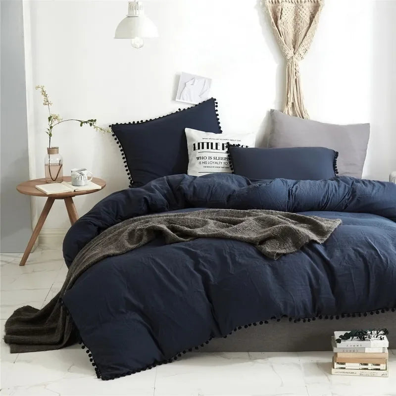A dark blue duvet cover set with a pom-pom trim displayed on a bed in a bright room.