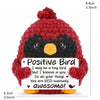 A handmade red crochet bird doll with a black face and an orange beak, featuring a positive message card.