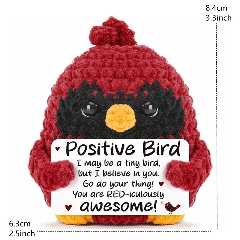 A handmade red crochet bird doll with a black face and an orange beak, featuring a positive message card.