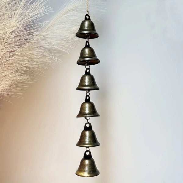 A vertical arrangement of small, antique bronze metal wind chimes hanging from a chain against a light background.