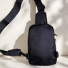 A black compact water-resistant sling bag is shown with its strap attached, viewed from the back.