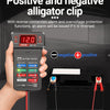 A hand holds a digital 12V battery tester with an LED display, connected to a car battery terminal.
