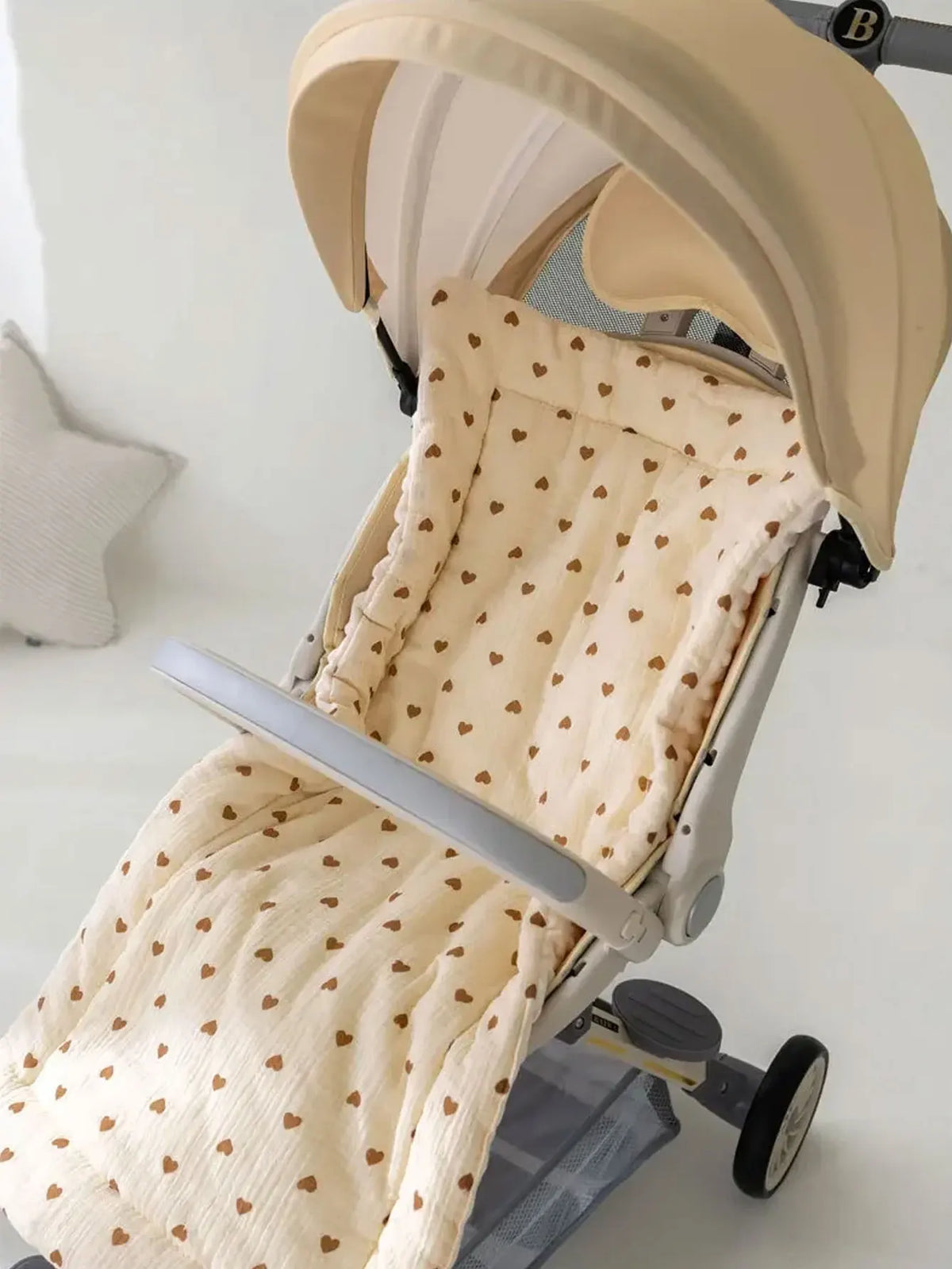 A beige stroller cushion with a pattern of small brown hearts is shown inside a light-colored stroller frame.
