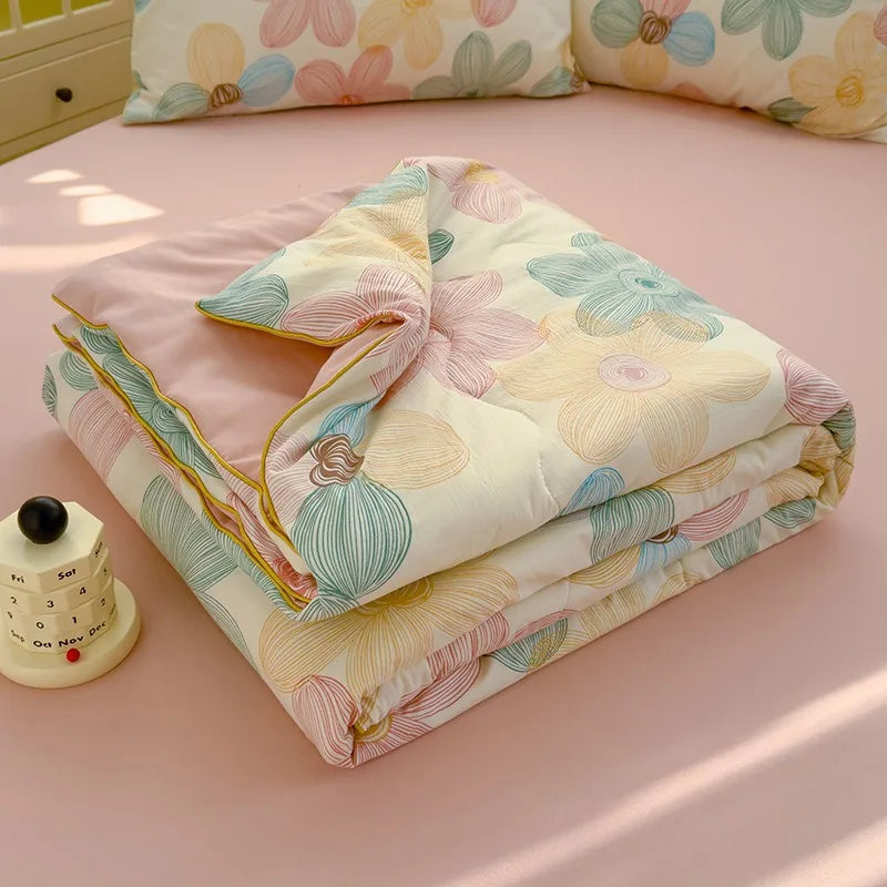 A folded lightweight washed cotton summer quilt with floral patterns in pink, yellow, and blue tones on a pink surface.