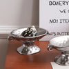 A silver ceramic soap dish with an elevated stand holds a small, patterned object on a wooden surface.