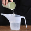 A hand pours liquid from a light green measuring cup into a clear plastic measuring jug.