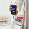 A hand holds a black portable digital luggage scale with an LCD display showing 0.045 kg, next to a white toilet.