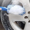 A hand is using a blue cleaning brush to wash and clean the surface of a silver car wheel.