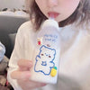 A person holds a white frosted water bottle featuring a blue outline of a bear and a strawberry, with a pink straw visible.