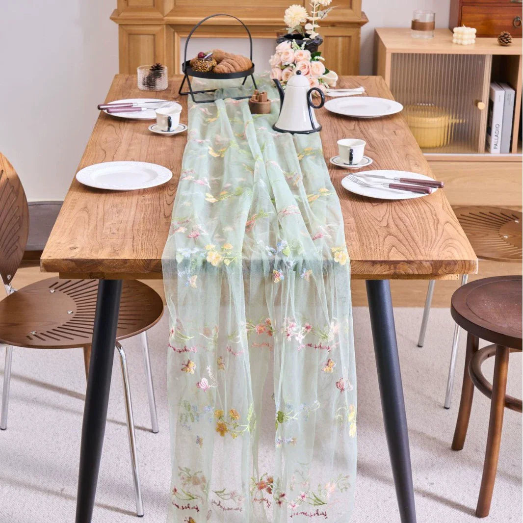 A sheer mint green tulle table runner with floral embroidery placed on a wooden dining table.