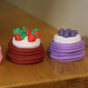 A colorful cake with strawberry and raspberry decorations is shown next to a purple cake on a wooden surface.