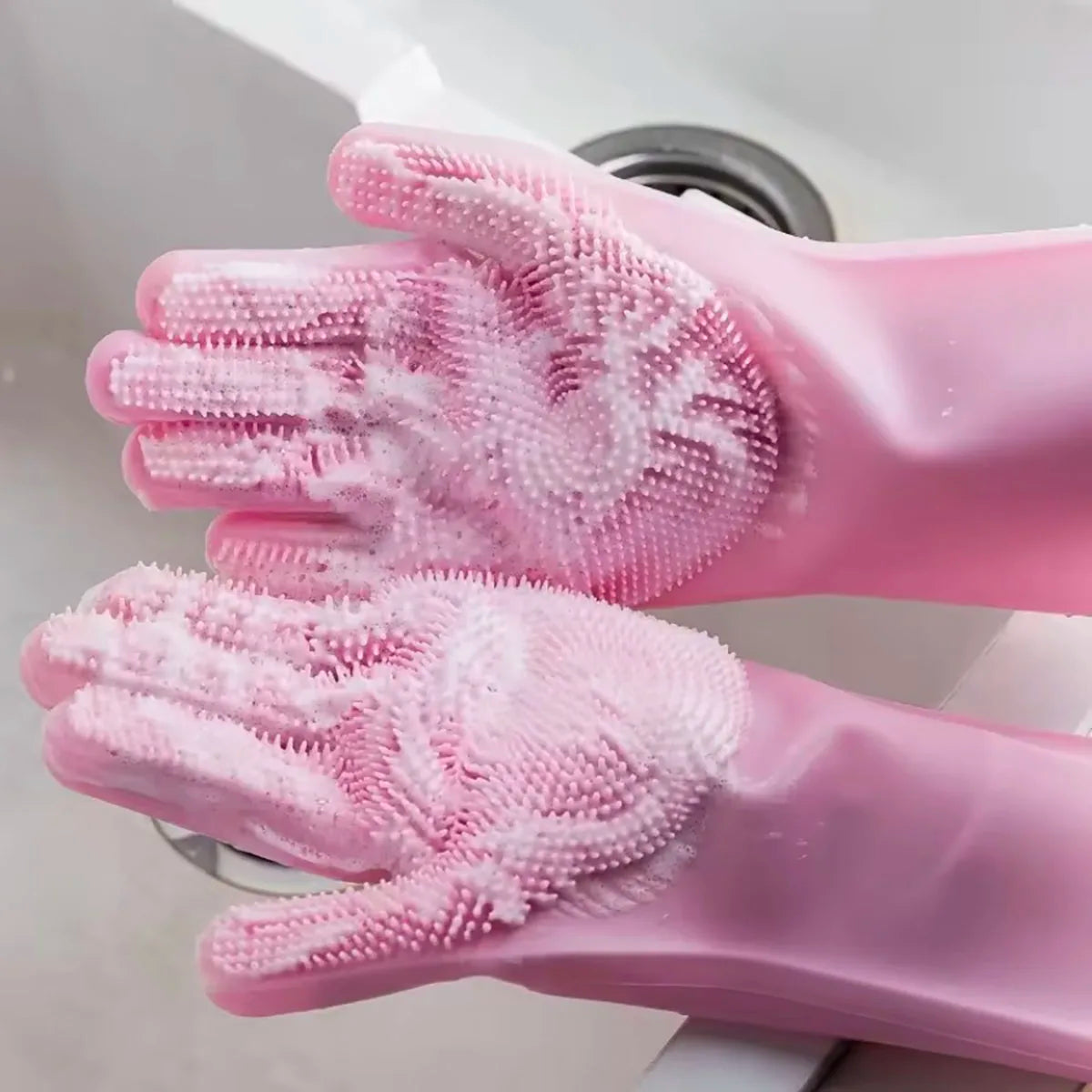 A pair of pink scrubber-textured dishwashing gloves are shown in a close-up against a white sink.