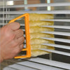 A hand is holding a yellow dusting brush with yellow fibers, positioned near white horizontal slats of a blind or vent.