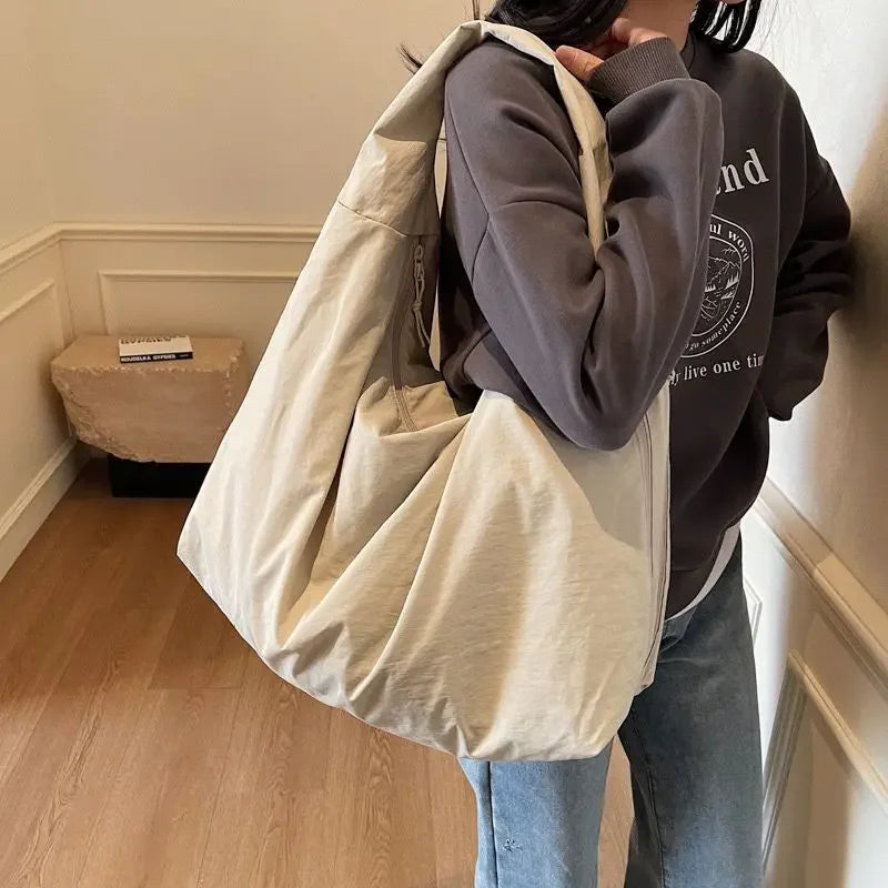 A person is holding a light beige nylon tote bag with a secure zipper closure against a white wall.