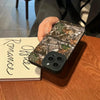 A hand holds a forest camouflage silicone phone case resting on a wooden table next to a notebook.